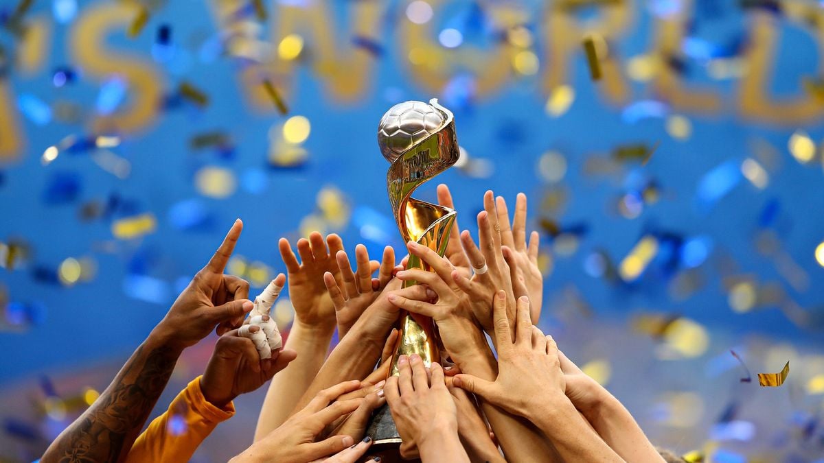 Multiple hands lift a golden soccer trophy together amidst falling blue and gold confetti, celebrating a victory against a blurred blue background.