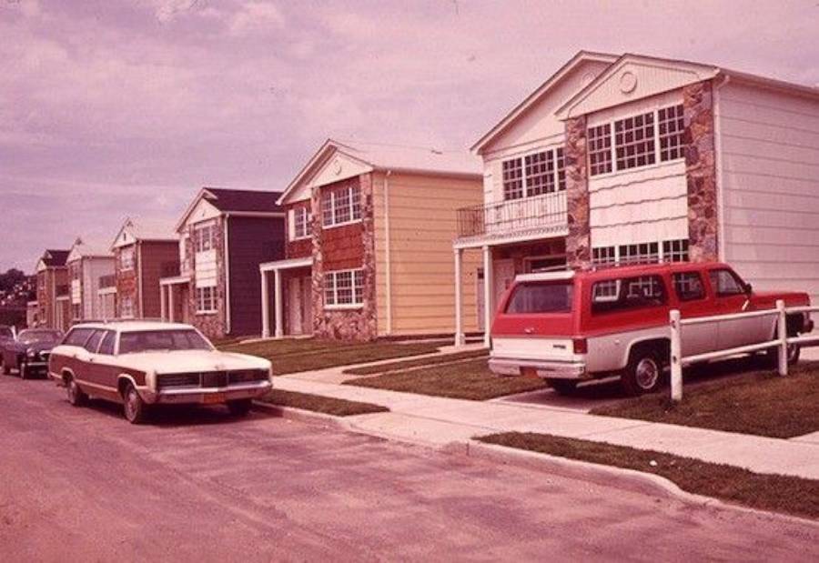 A row of suburban houses with boxy architecture and large windows, parked cars from the 1970s era line the street and driveways under a cloudy sky.