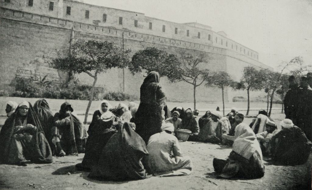 A black and white photo showing a group of people, many wearing cloaks or head coverings, sitting and standing in a circle outdoors near a large stone wall lined with trees.