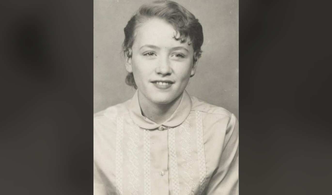 A young woman with short, wavy hair smiles slightly at the camera. She is wearing a buttoned blouse with lace details. The photo is black and white and has a vintage look.