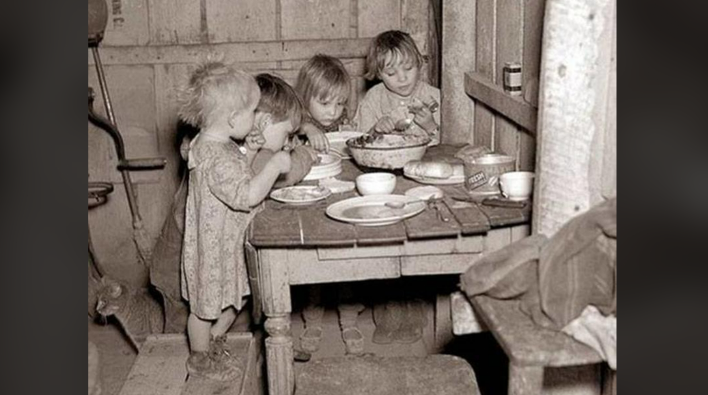 Four young children in worn clothing eat a meal together at a small wooden table in a rustic, shabby room. The table holds dishes, bread, and a bowl of food. The setting appears modest and old-fashioned.