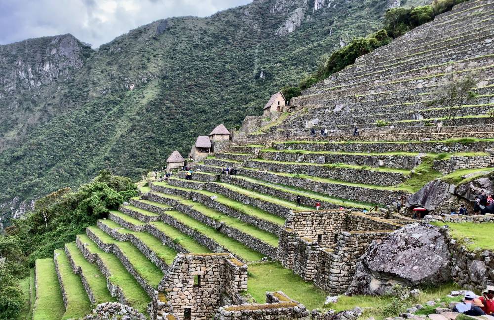 Stone terraces and ancient buildings of Machu Picchu rise along a green mountainside, with lush vegetation and misty mountains in the background. Tourists are visible exploring the site.