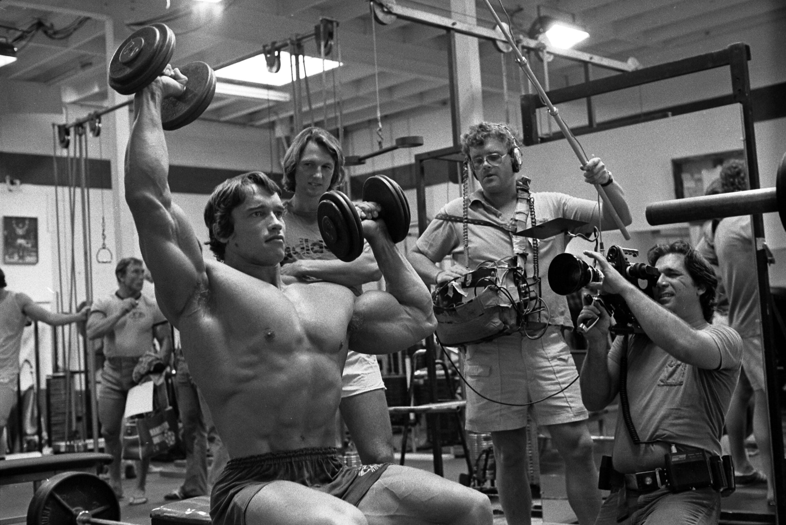 A muscular man lifts dumbbells overhead in a gym while three men, one with a camera and another with audio equipment, film him during his workout. The gym is busy with various equipment visible in the background.