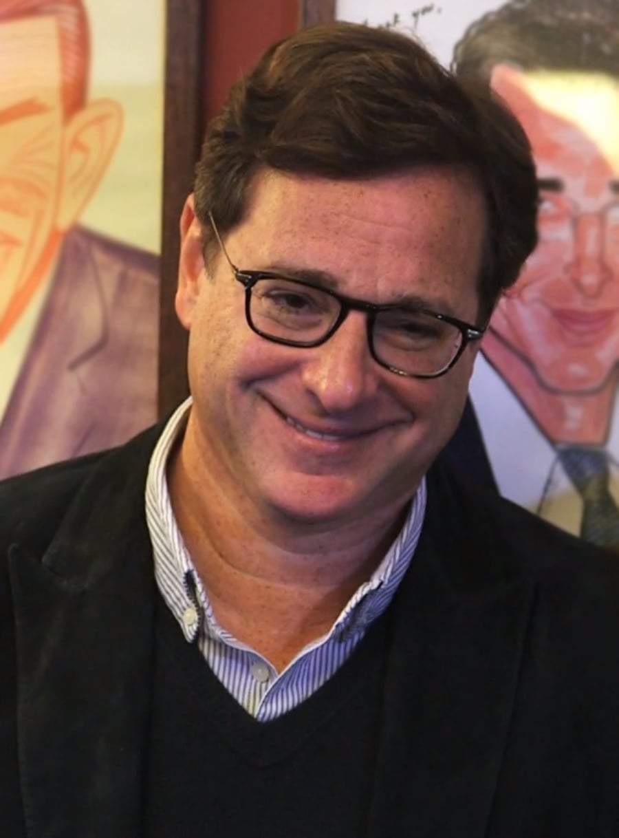 A man with brown hair and glasses smiles while wearing a dark blazer over a striped shirt and sweater, standing in front of colorful portraits.