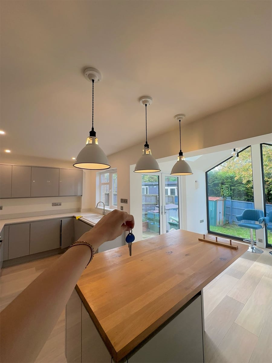 A hand holds a blue key over a modern kitchen island with a wooden countertop, pendant lights above, and large windows overlooking a garden with outdoor seating.
