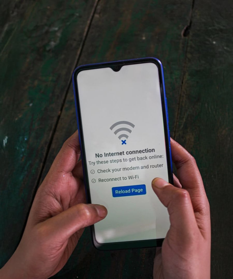 A person holding a smartphone displaying a "No Internet connection" error message, suggesting to check modem and router, reconnect to Wi-Fi, and showing a "Reload Page" button.