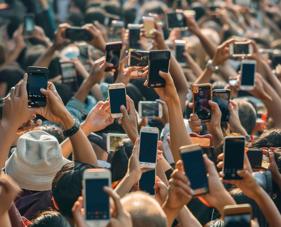 A large crowd of people hold up smartphones, taking photos or recording videos. The scene is densely packed, and almost every person has a phone raised, capturing something out of frame.