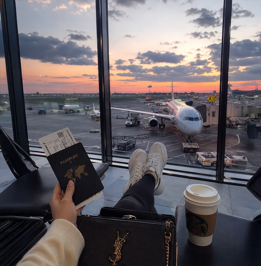 A person relaxes at an airport gate with legs crossed, holding a passport and boarding pass. A coffee cup and purse rest nearby, while an airplane sits by the window at sunset.