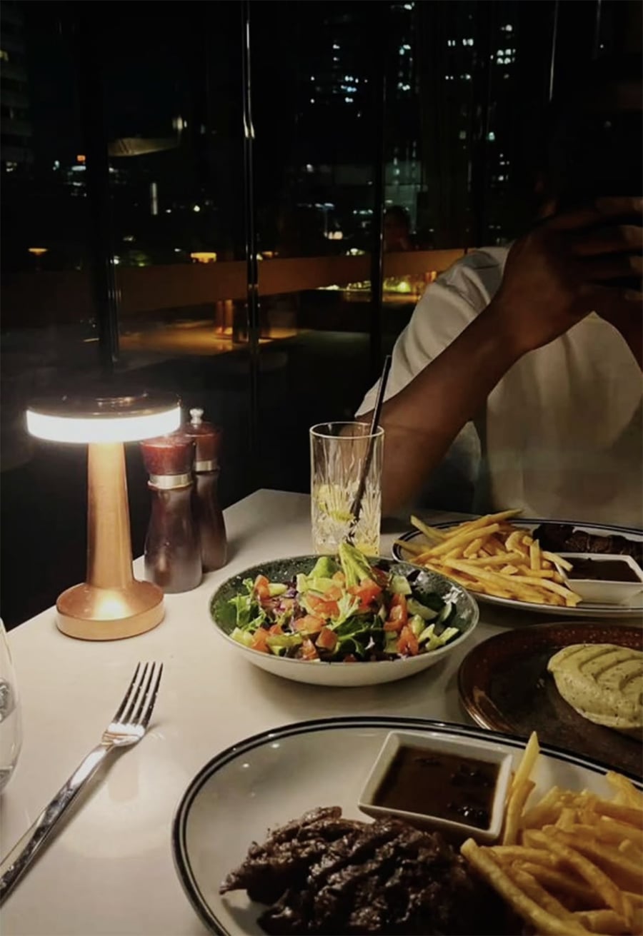 A dimly lit restaurant table with plates of steak, fries, salad, a tall drink, and a small lamp. A person in a white shirt sits at the table, city lights visible through the window in the background.