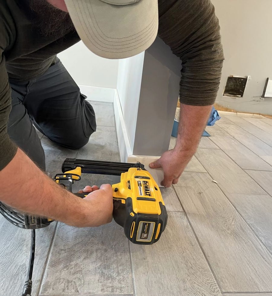 A person kneels on a tiled floor using a yellow DeWalt nail gun to attach white baseboard trim to a wall in a home interior under renovation.