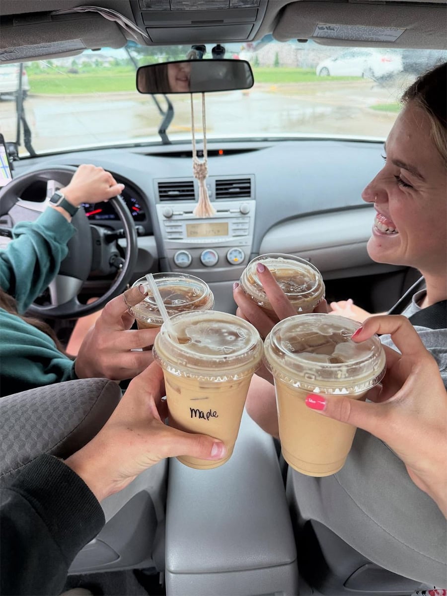 Four people inside a car holding iced coffee drinks together in a toast. One person is driving, while a smiling passenger sits in the front seat. It is raining outside, and the drinks have clear lids and straws.