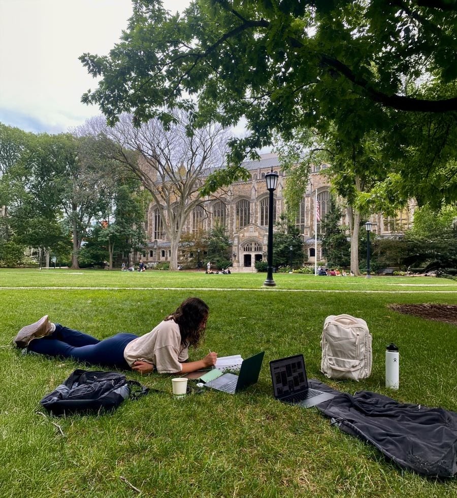 A person lies on grass under a tree, working on a laptop with notebooks, a cup, backpack, and water bottle nearby. In the background, a large, historic building is visible across a green lawn.