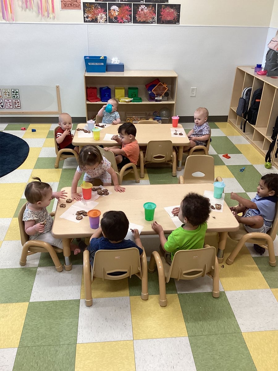 A group of toddlers sits at two small tables in a classroom, eating snacks and drinking from colorful cups. The room has shelves with toys and backpacks, and the floor is green and yellow checkered.