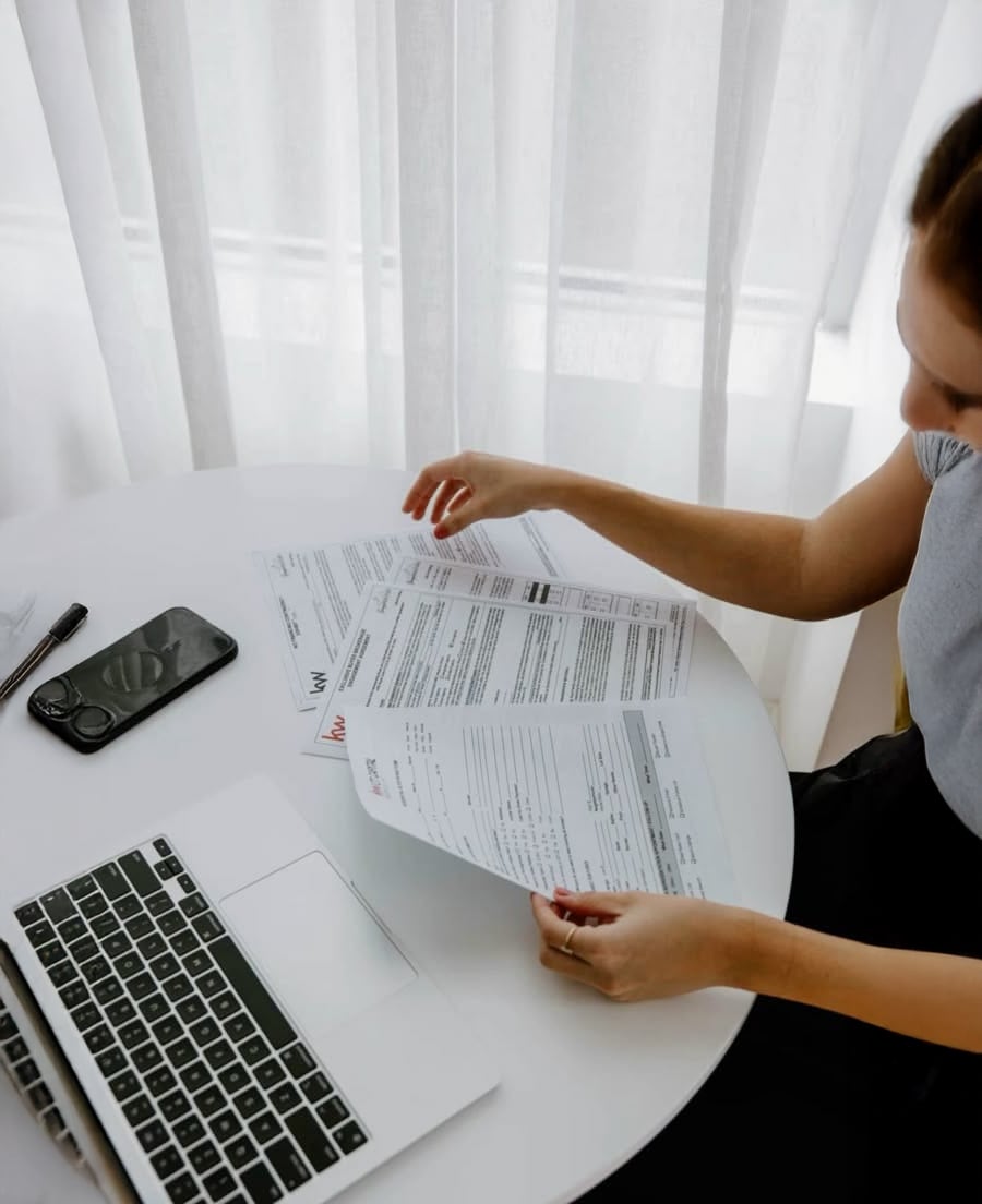 A person sits at a round white table, reviewing documents with text, alongside an open laptop, a smartphone, and a pen, near a window with sheer white curtains.