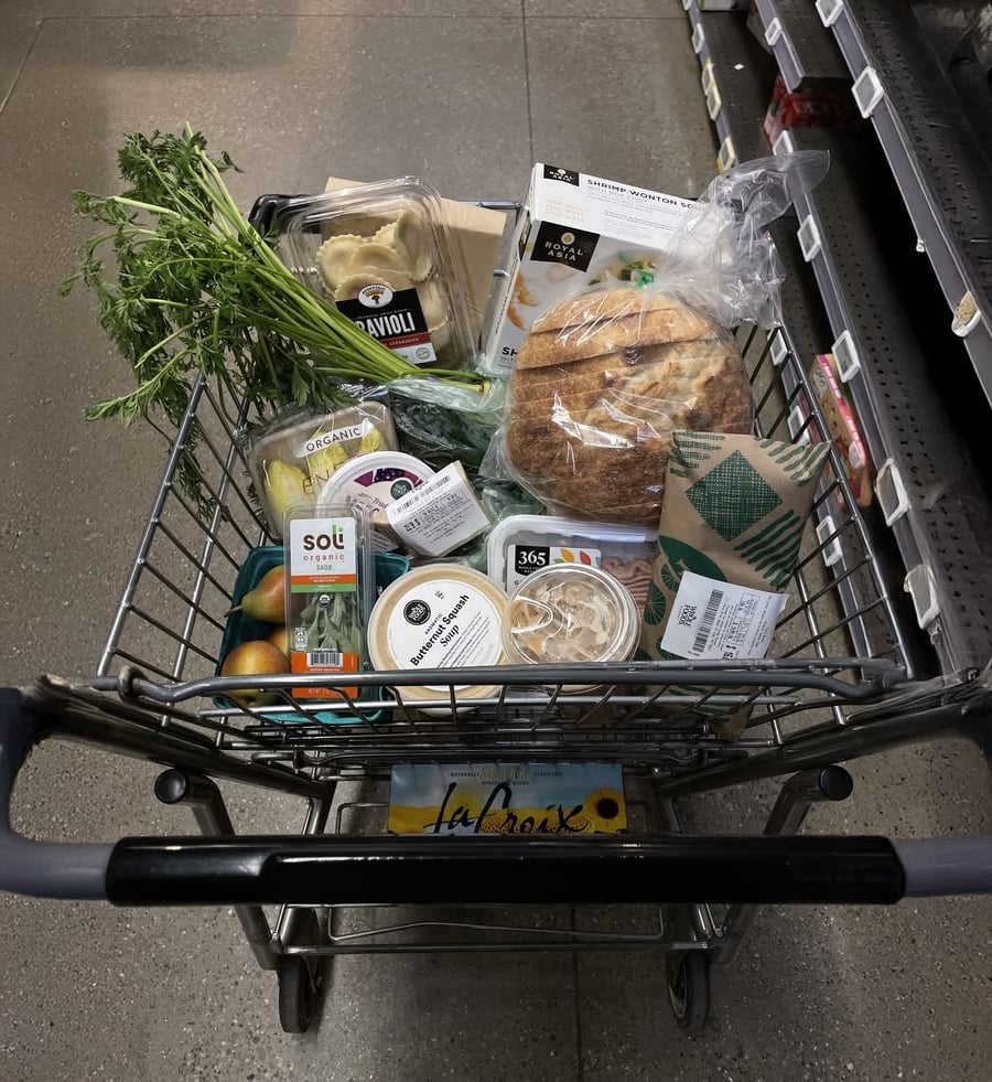 A grocery cart filled with fresh parsley, ravioli, artisan bread, salad greens, tomatoes, hummus, cheese, a box of eggs, and other packaged food items in a supermarket aisle.