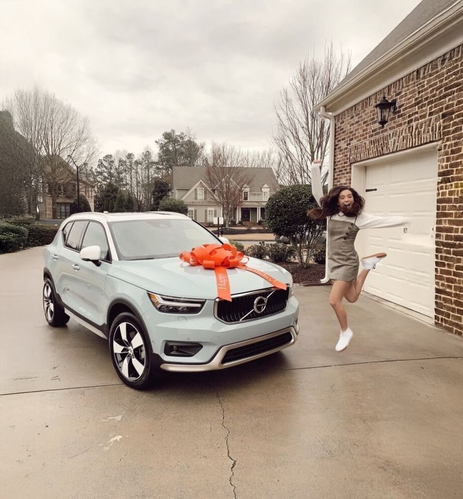 A young woman jumps excitedly next to a light blue Volvo SUV with a large red bow on the hood, parked in a driveway in front of a brick house on a cloudy day.