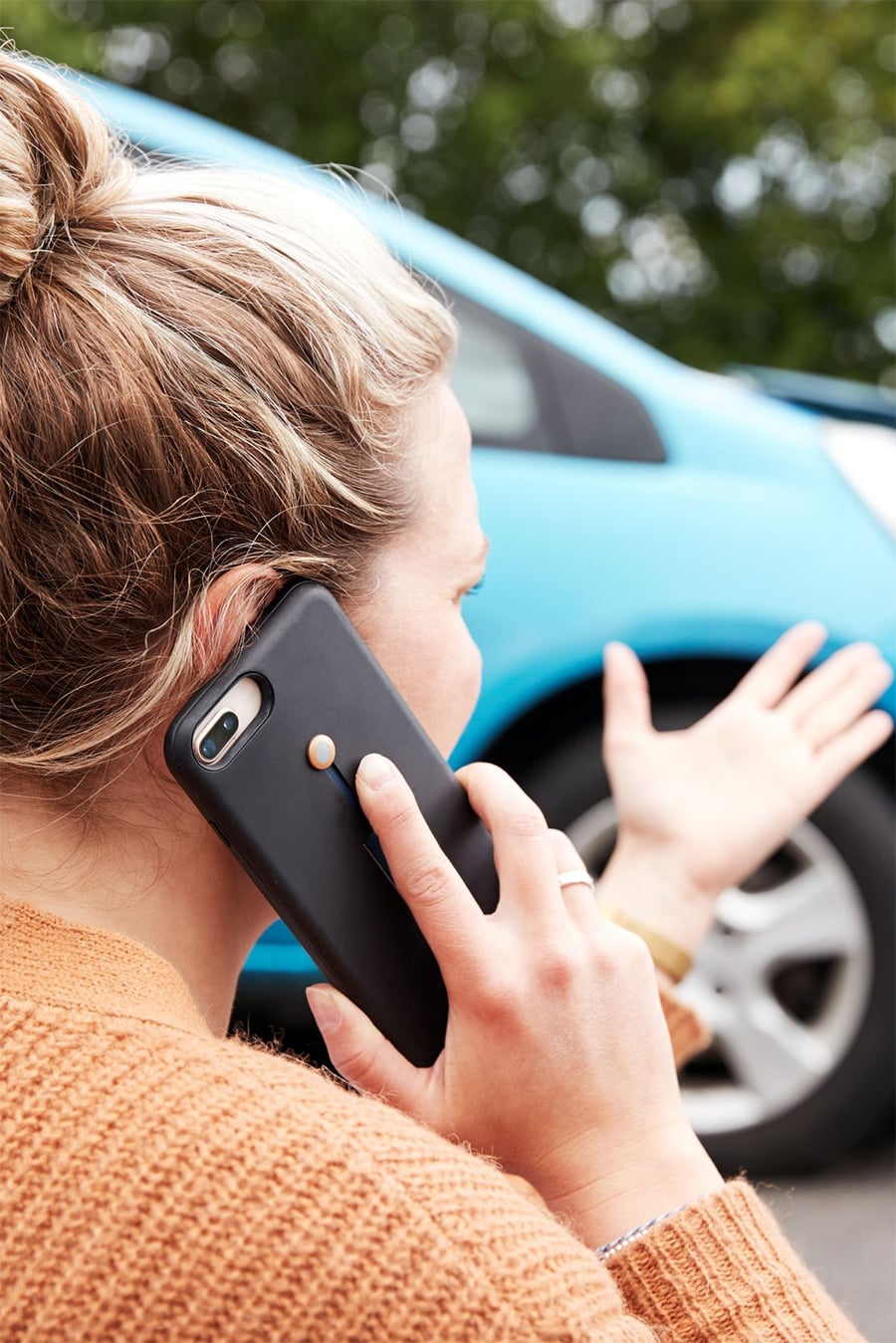 A woman with blonde hair in a bun speaks on her smartphone while gesturing with her hand, standing outside near a blue car. She wears an orange sweater and appears concerned.