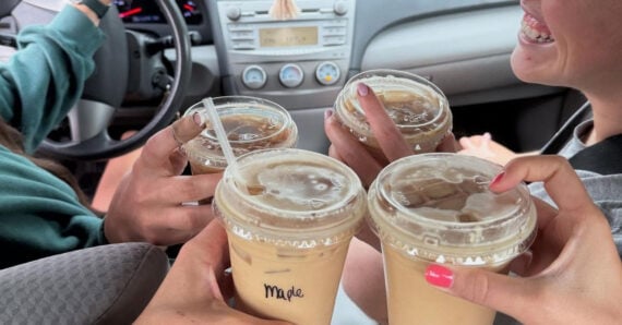 Four people in a car clink their iced coffee drinks together, celebrating. One cup has the name "Myle" written on it. The mood appears cheerful, with one person smiling brightly.