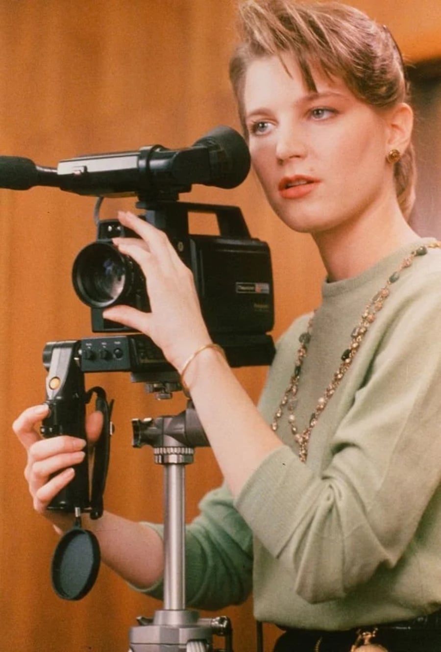 A woman with blonde hair, wearing a light green sweater and a necklace, is operating a vintage video camera mounted on a tripod indoors.