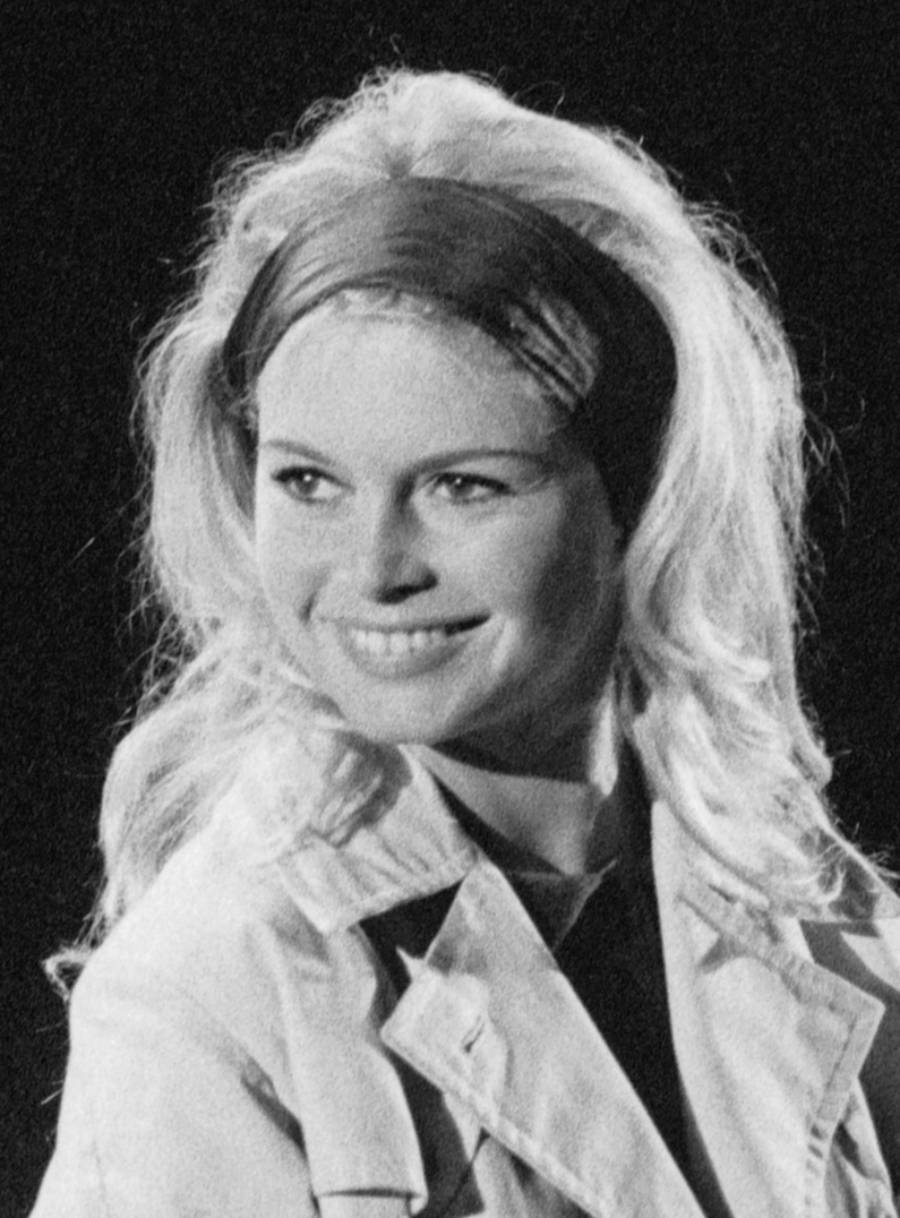 A woman with long, wavy blonde hair and a headband smiles while looking to the side. She is wearing a light-colored jacket over a dark top, and the background is dark.