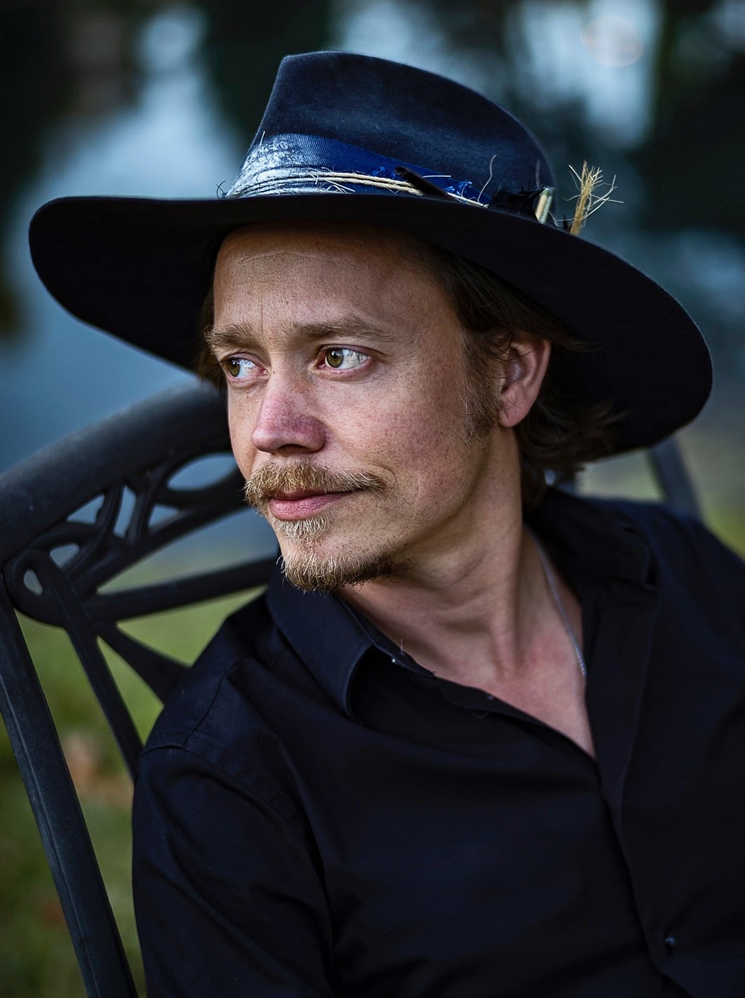 A man with light brown hair and a goatee sits outdoors on a chair, wearing a dark wide-brimmed hat with a feather and a black shirt, looking thoughtfully to the side.
