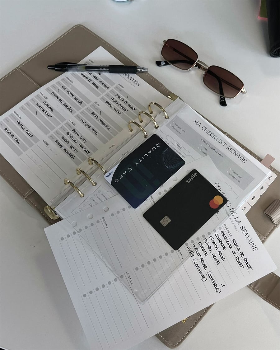 A beige planner open on a desk shows weekly checklists, a pen, two credit cards, a list, and sunglasses. The pages are organized in rings and include handwritten notes and tasks.
