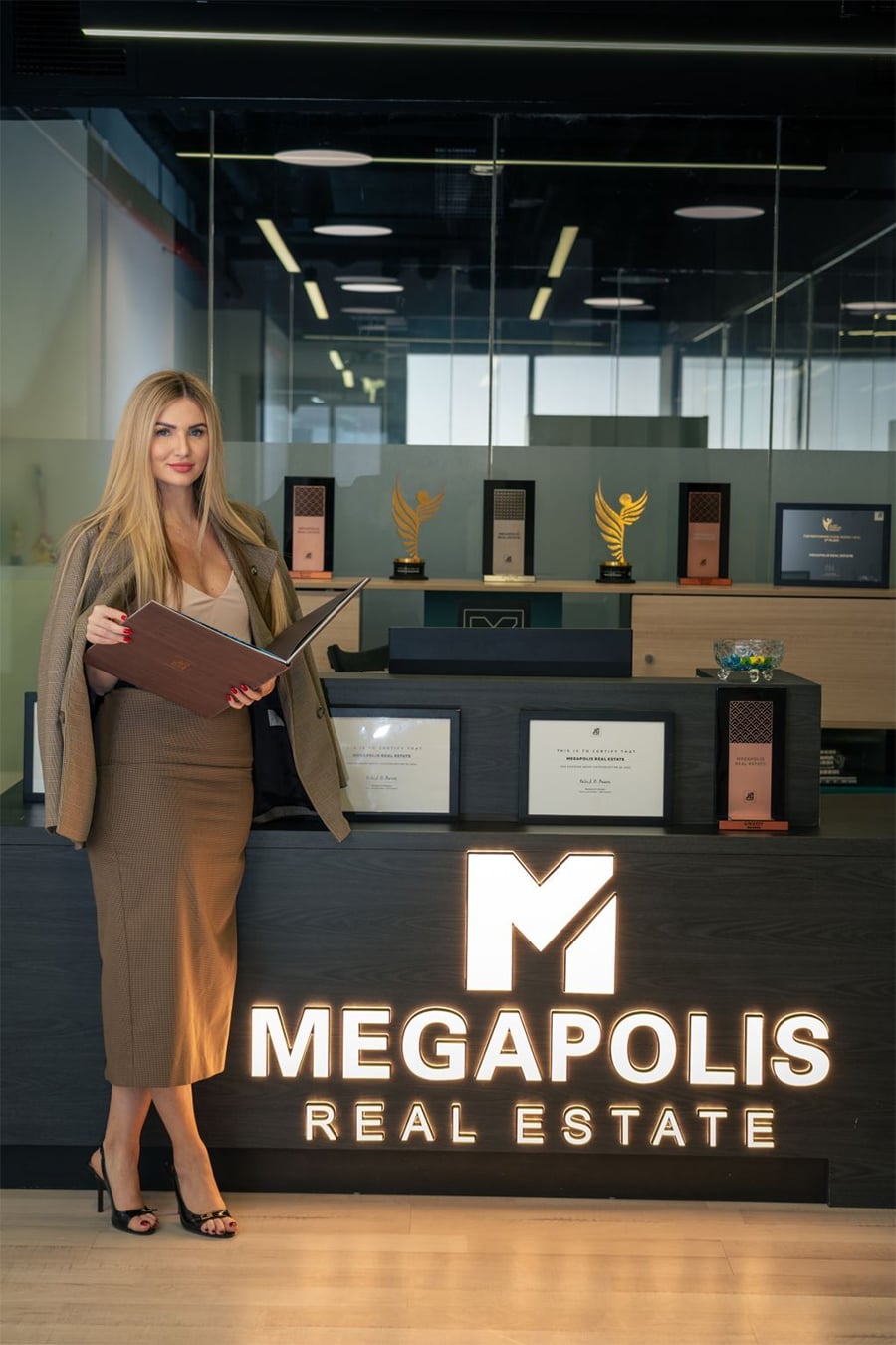 A woman in business attire stands next to a lit "MEGAPOLIS REAL ESTATE" sign, holding an open folder. Several awards and certificates are displayed on a counter behind her in a modern office setting.