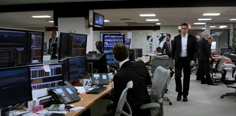 A modern office with multiple people working at desks filled with computer monitors displaying financial data. One man in a suit is walking while others are seated and focused on their screens. The atmosphere is busy and professional.