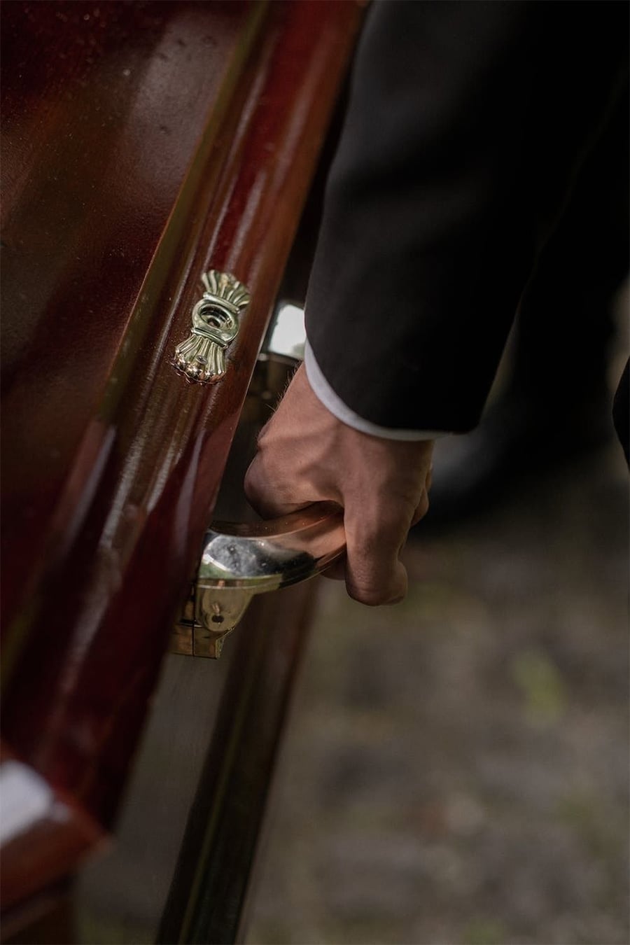 A close-up of a person in a dark suit holding the silver handle of a polished wooden coffin during a funeral.