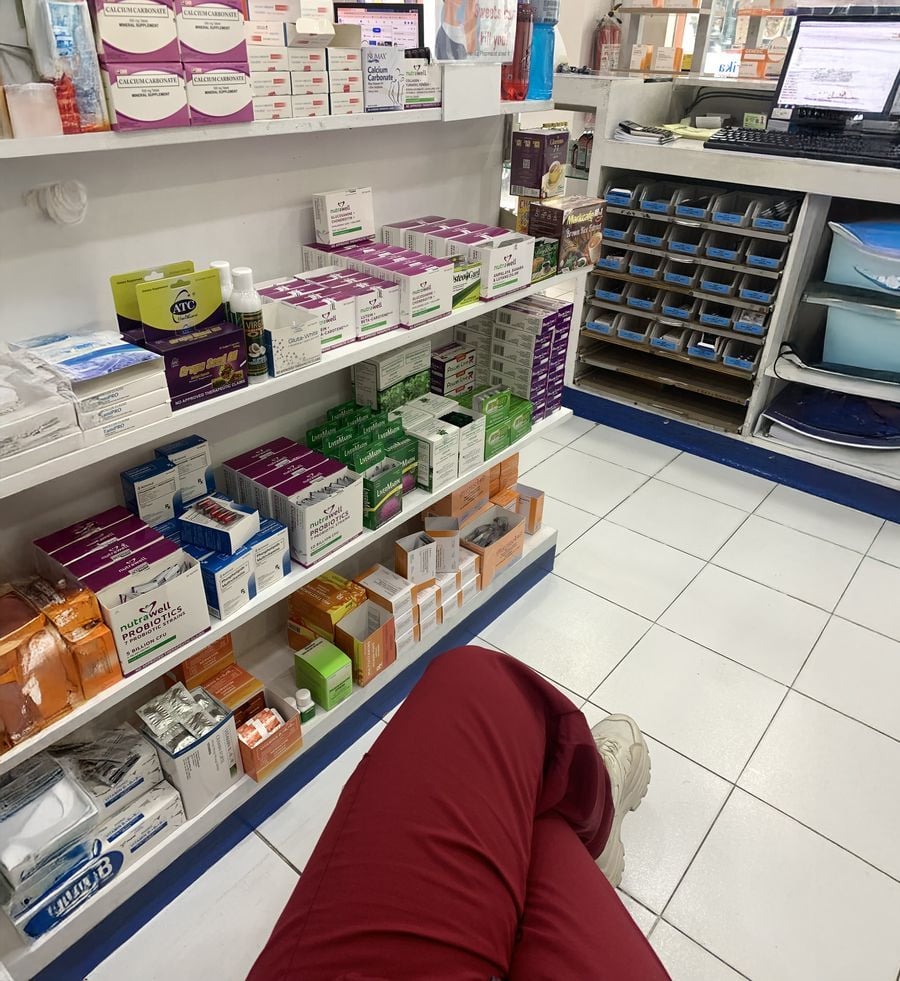 A person in red pants sits in a pharmacy or medical supply room, with shelves of medication boxes, ointments, and health products. A computer workstation and blue storage bins are visible in the background.