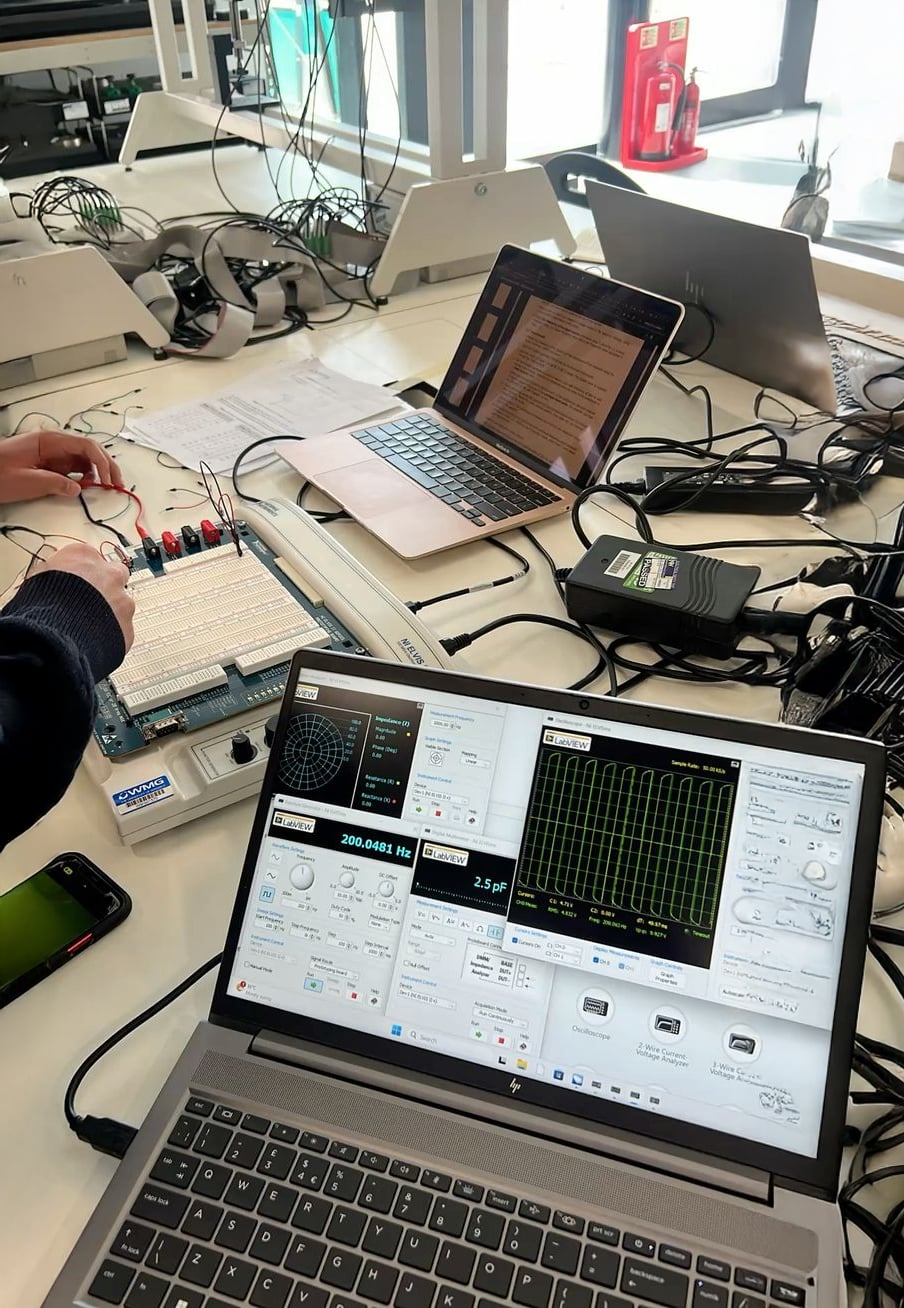 A workspace with laptops, an oscilloscope display on one screen, a breadboard with wires, a phone, and various cables. Someone's hands are adjusting the breadboard in an electronics lab setting.