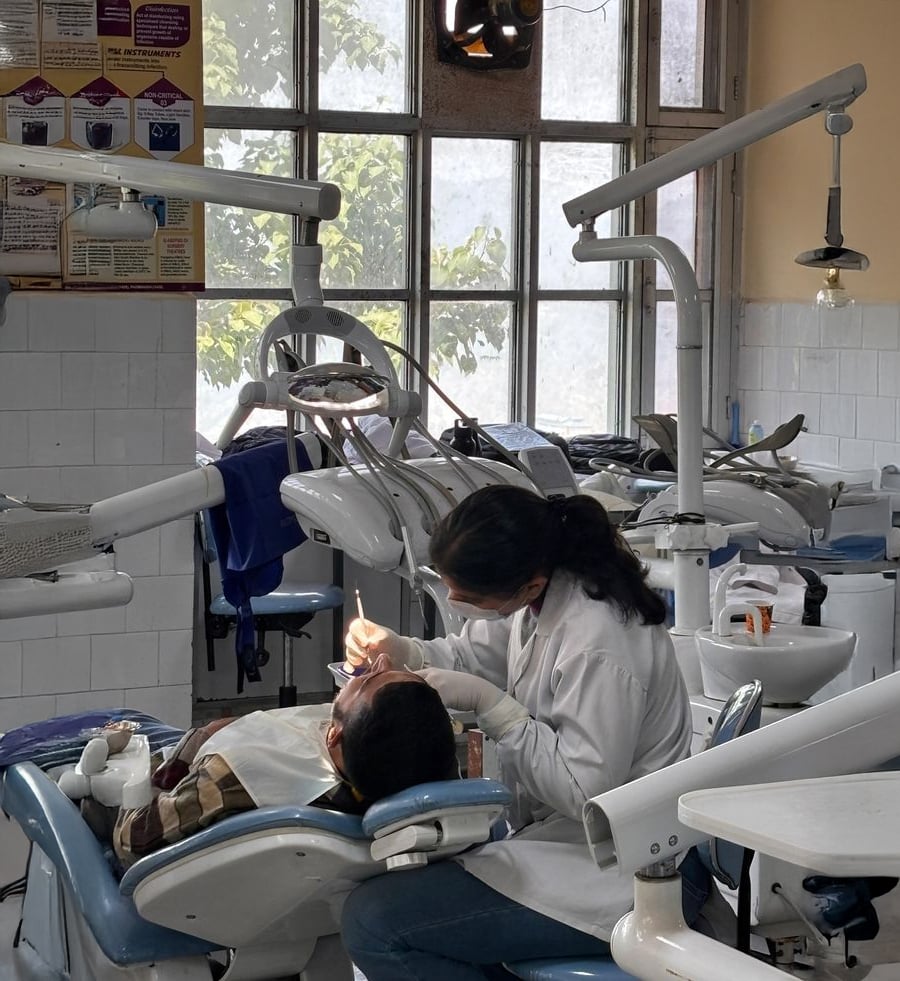 A dentist wearing a white coat and mask examines a patient’s teeth in a dental clinic with dental equipment and large windows in the background.