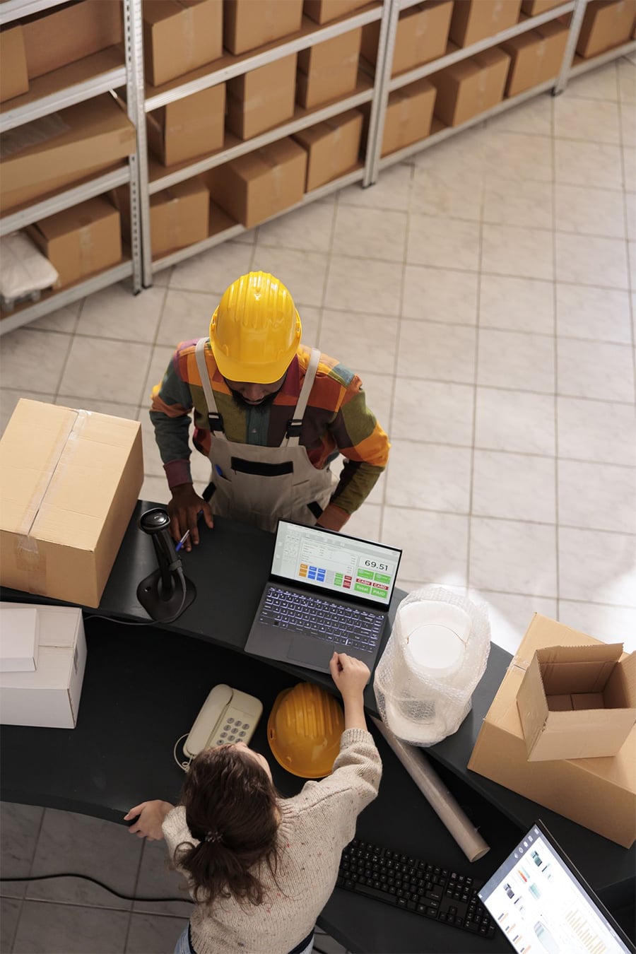 A warehouse worker in a yellow hard hat stands at a counter with boxes, while a woman points at a laptop displaying charts. Shelves with packages are visible in the background.