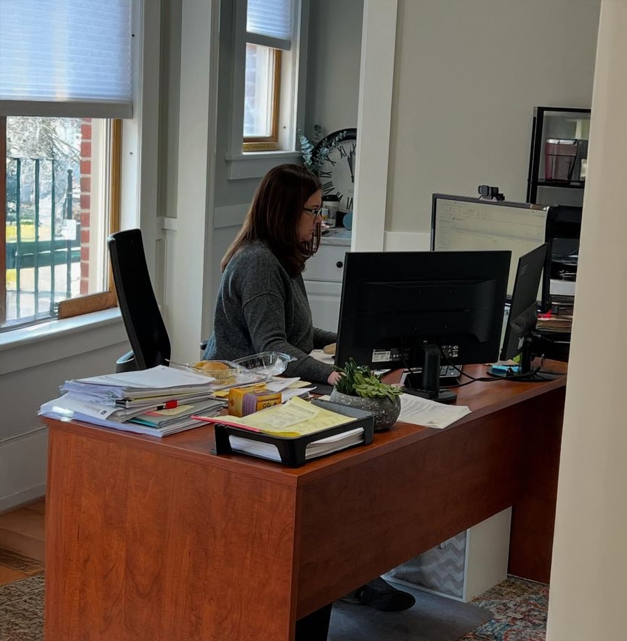 A woman with dark hair and glasses is working at a computer in a bright office with large windows. Papers, folders, and office supplies are scattered on the wooden desk.