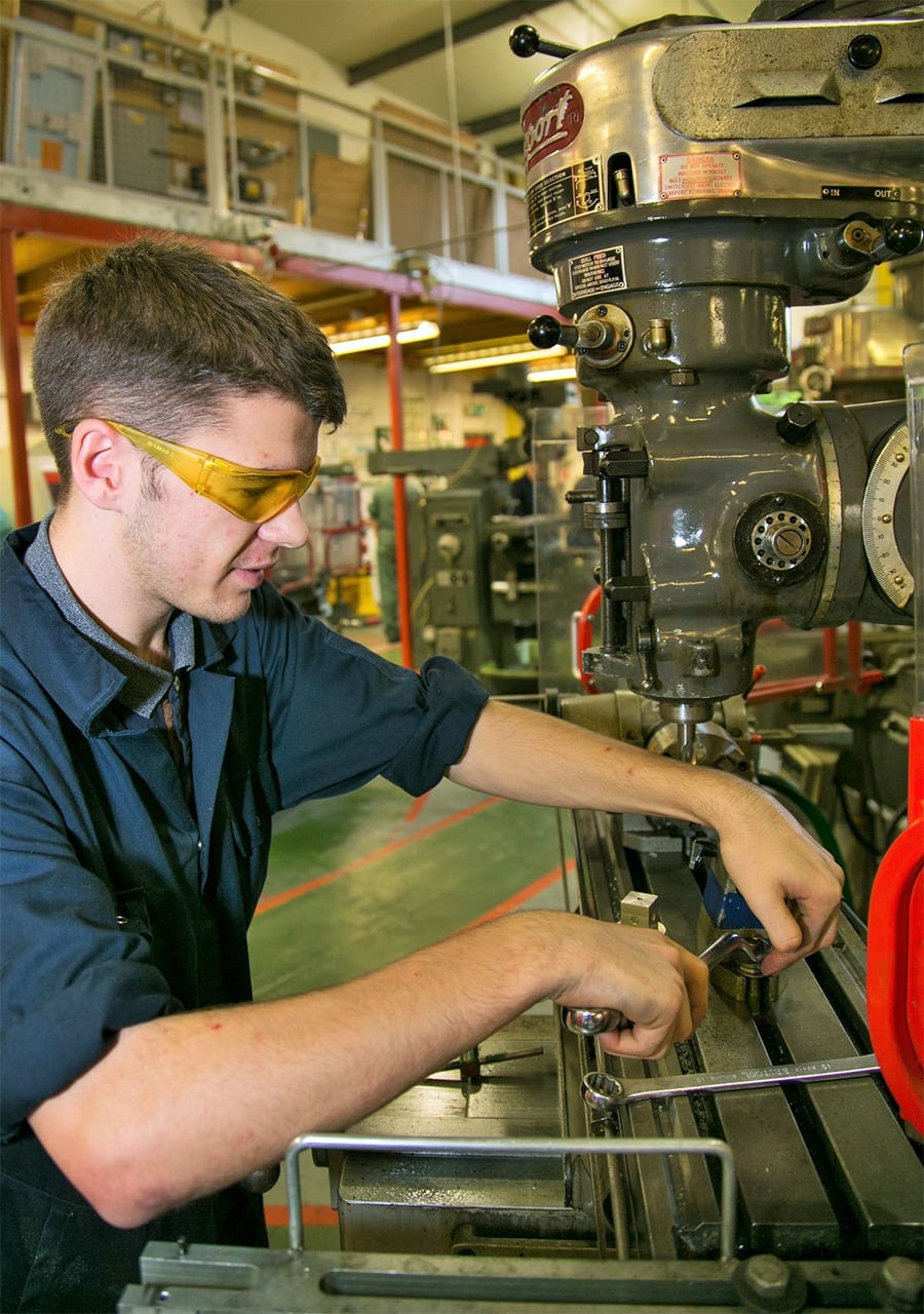 A man wearing safety glasses and work clothes operates a large industrial drill press in a workshop, adjusting a piece of metal clamped to the machine.