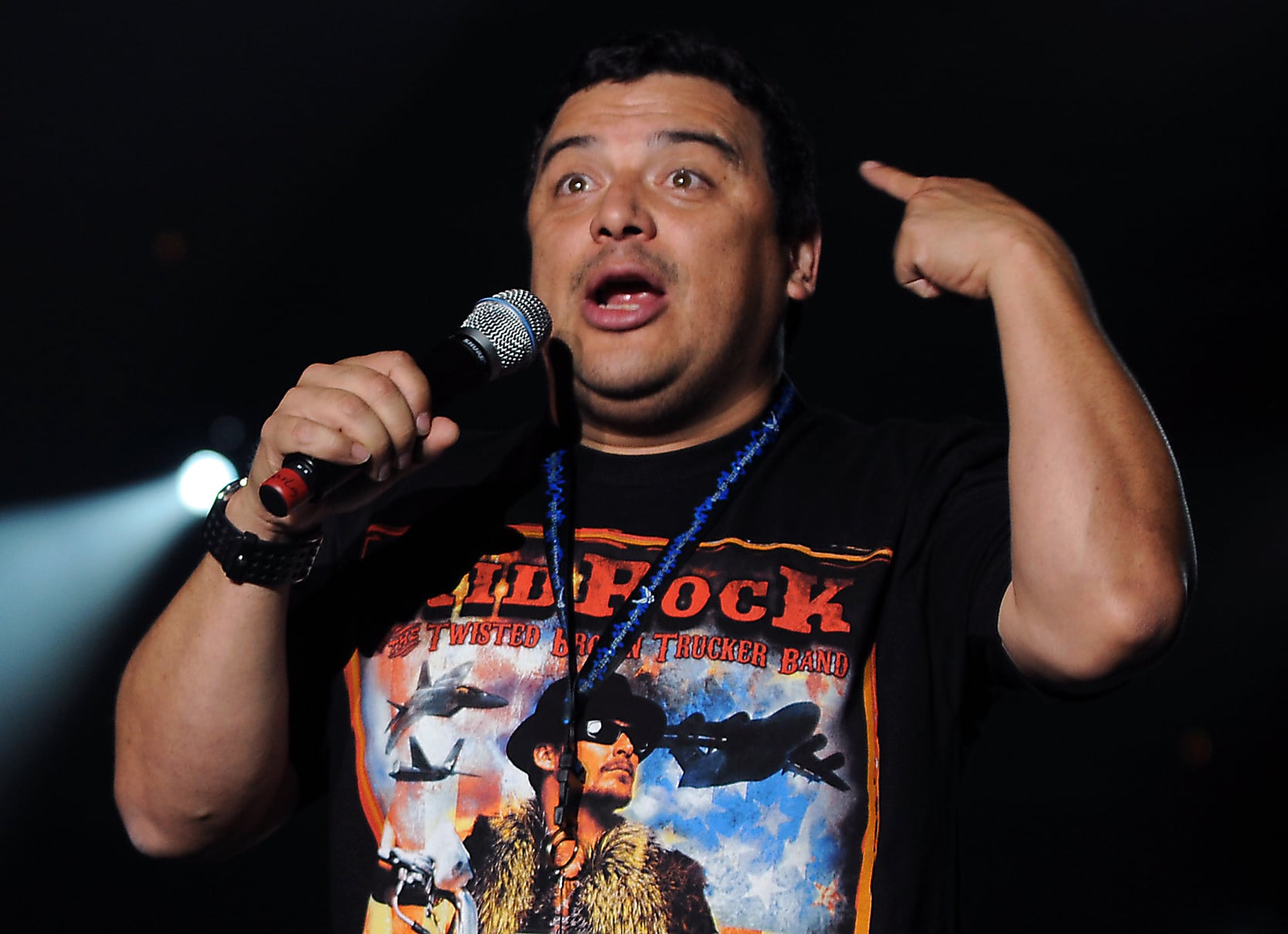 A comedian on stage holds a microphone and gestures with one hand toward his head. He wears a black T-shirt with a colorful graphic and appears to be speaking animatedly, with a spotlight shining behind him.