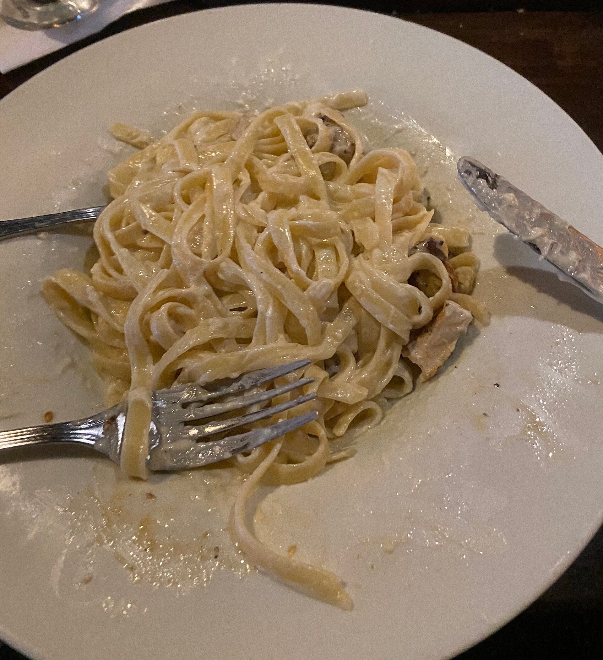 A partially eaten plate of fettuccine Alfredo with pieces of chicken, a fork, and a knife resting on the white plate. The creamy sauce is spread around the pasta and plate.