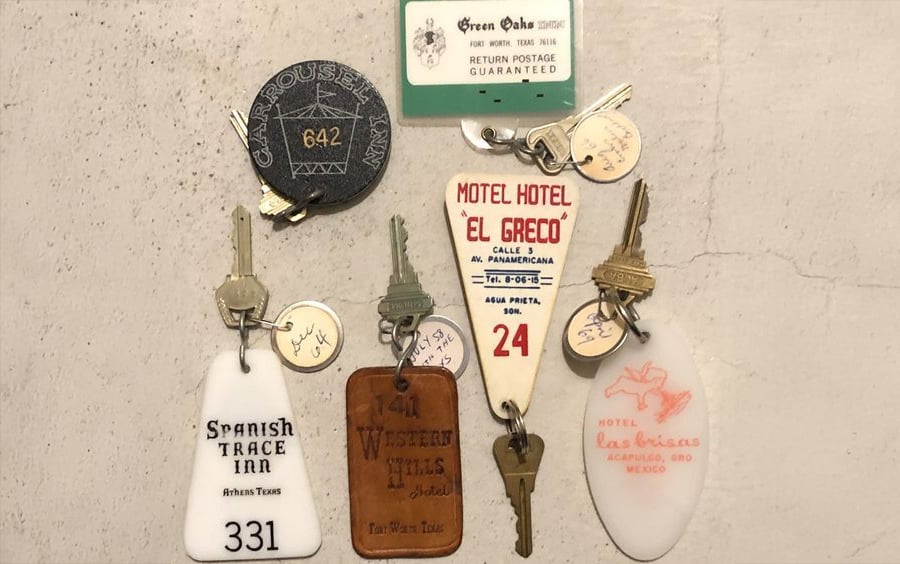A display of six vintage motel room keys with labeled keychains, featuring hotel names, addresses, and room numbers in various shapes and colors, mounted on a white wall.