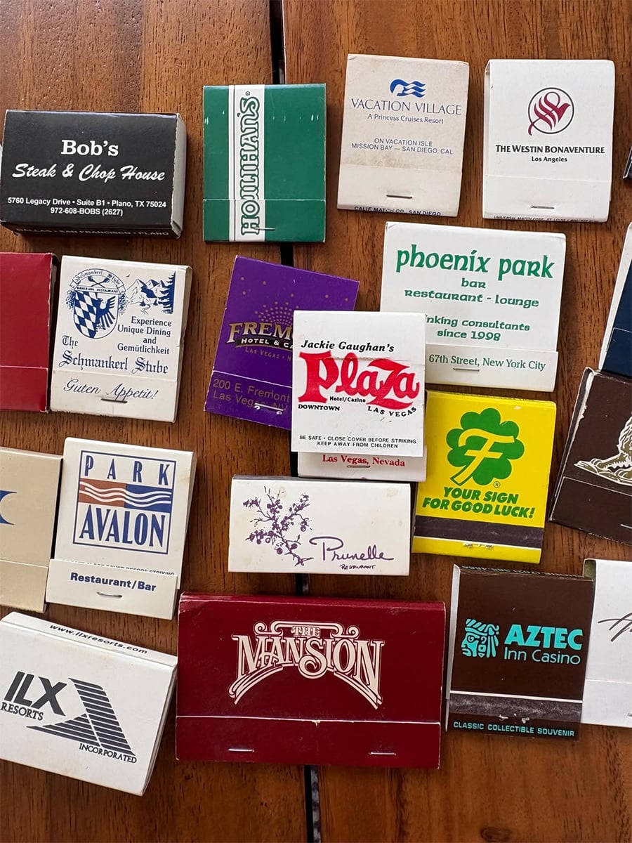 A collection of colorful vintage matchbooks from various hotels, casinos, and restaurants is spread out on a wooden surface. The matchbooks display different logos, names, and designs.