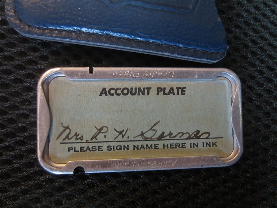 A metal account plate with a paper insert labeled "ACCOUNT PLATE" and "PLEASE SIGN NAME HERE IN INK," displaying a handwritten signature. A textured black surface and part of a blue leather item are visible.