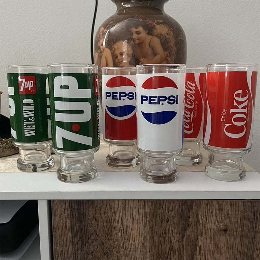 Six drinking glasses turned upside down, each decorated with retro soft drink logos for 7UP, Pepsi, and Coca-Cola, displayed on a wooden surface with a decorative lamp and painting in the background.