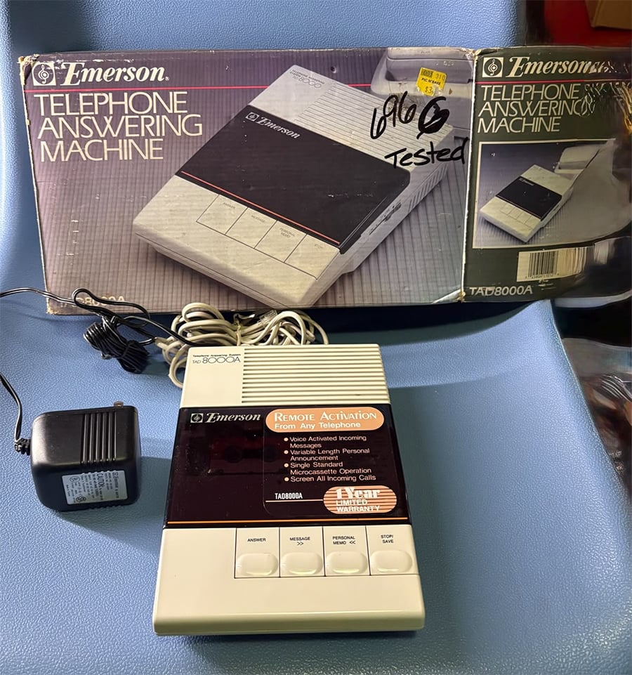 An Emerson telephone answering machine, model TAB8000A, is shown with its original box, power adapter, and cords on a blue surface. The box displays an image and description of the machine.