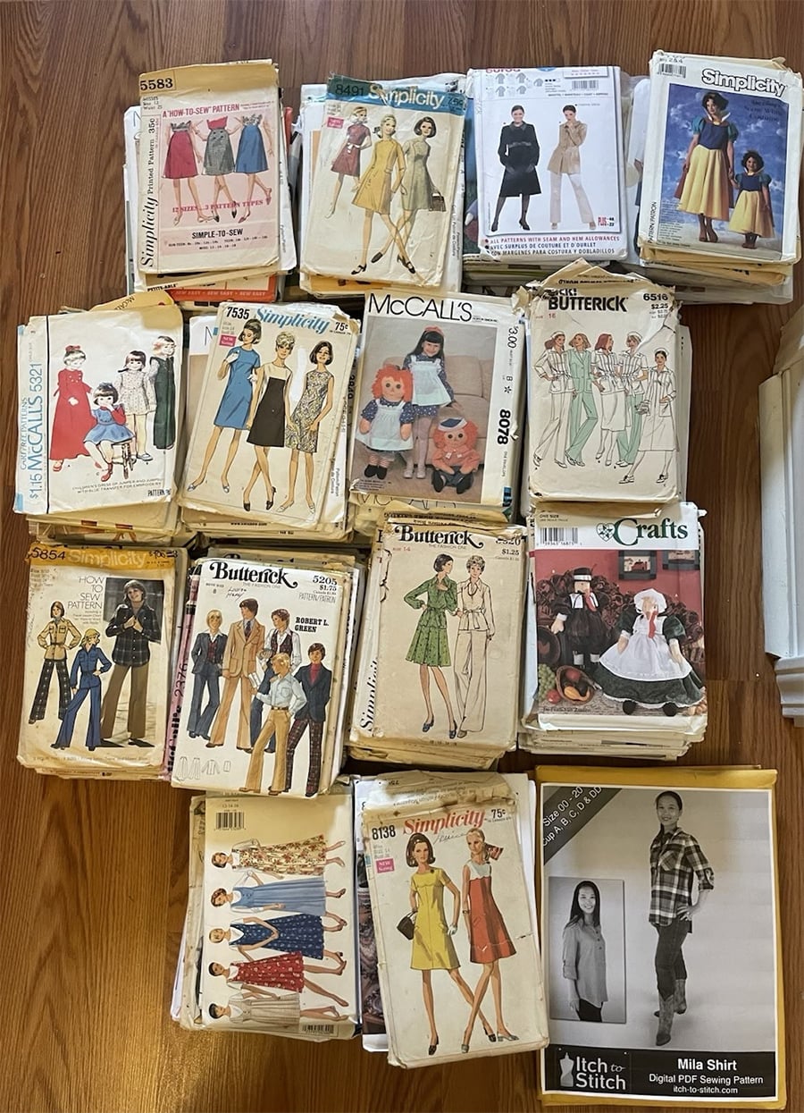 A collection of vintage sewing pattern envelopes arranged on a wooden floor, featuring illustrated women and girls in various outfits including dresses, coats, suits, and pants from brands like McCall’s, Butterick, Simplicity, and more.
