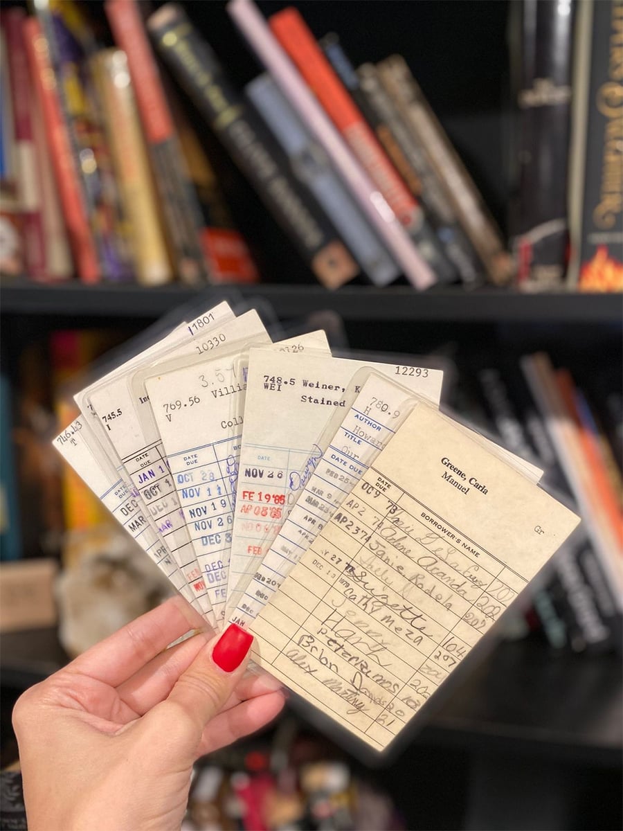 A hand with red nail polish holds several vintage library checkout cards, each stamped with dates, in front of a blurred bookshelf filled with assorted books.