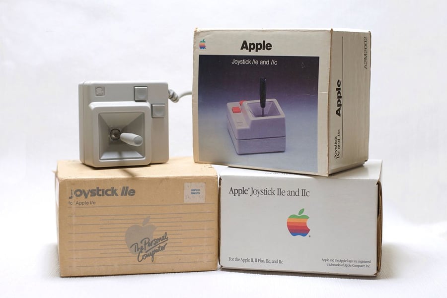 A vintage Apple joystick with a gray base and black stick is displayed in front of three stacked boxes labeled "Apple Joystick IIe and IIc," featuring old Apple branding and logos.