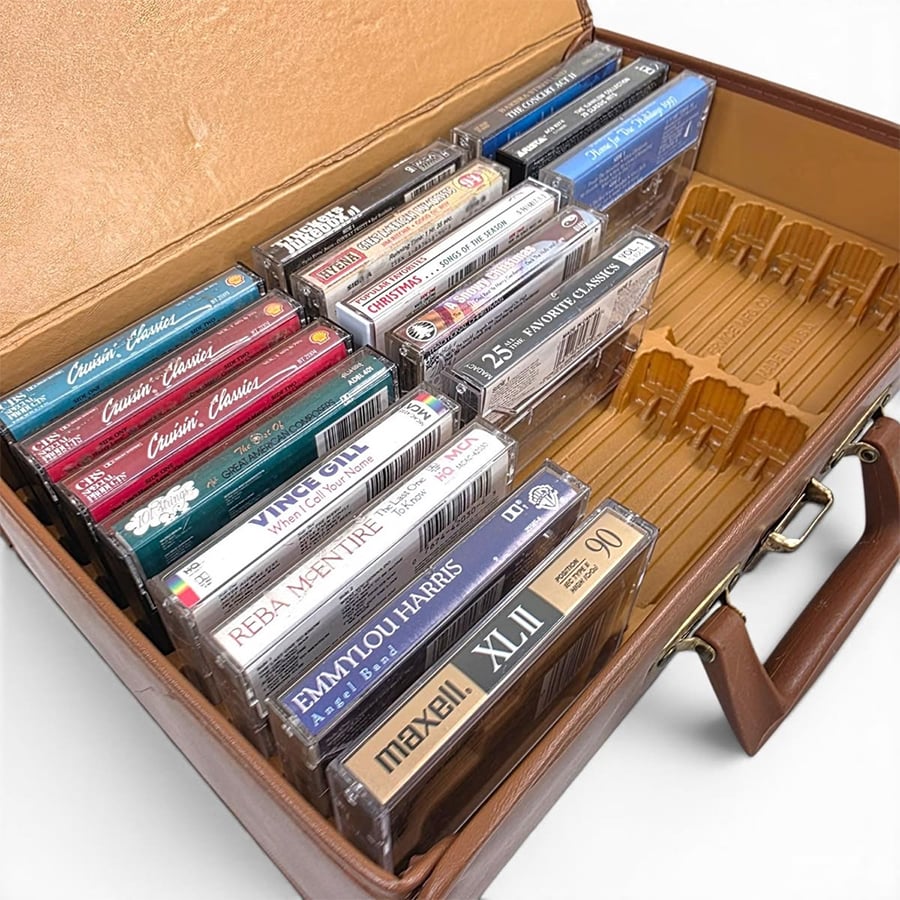 A brown briefcase holds several cassette tapes in plastic cases, organized upright in rows. Some cassette labels are visible, featuring various music artists and genres. The briefcase has additional empty slots for more tapes.
