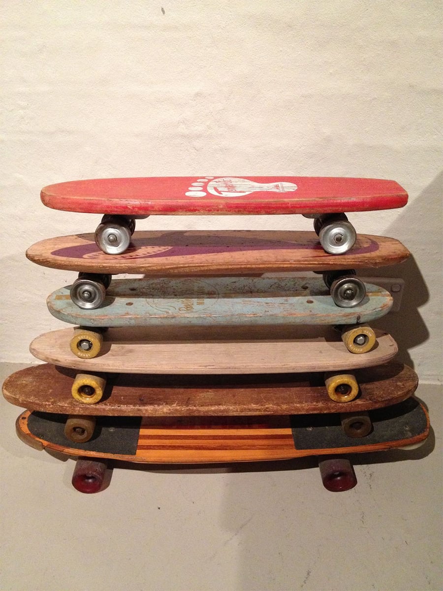 Five vintage skateboards are stacked horizontally against a beige wall, each with different colored decks and wheels, showing signs of wear and age.