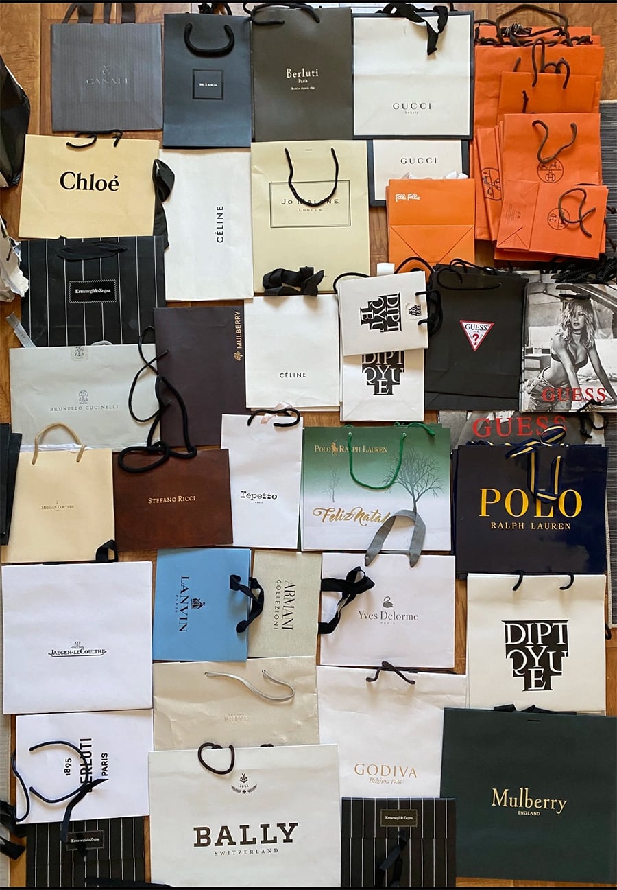 A variety of branded shopping bags are laid out on a flat surface, including labels such as Gucci, Prada, Polo Ralph Lauren, Godiva, Bally, Guess, Chloé, Céline, Mulberry, and Diptyque, among others.