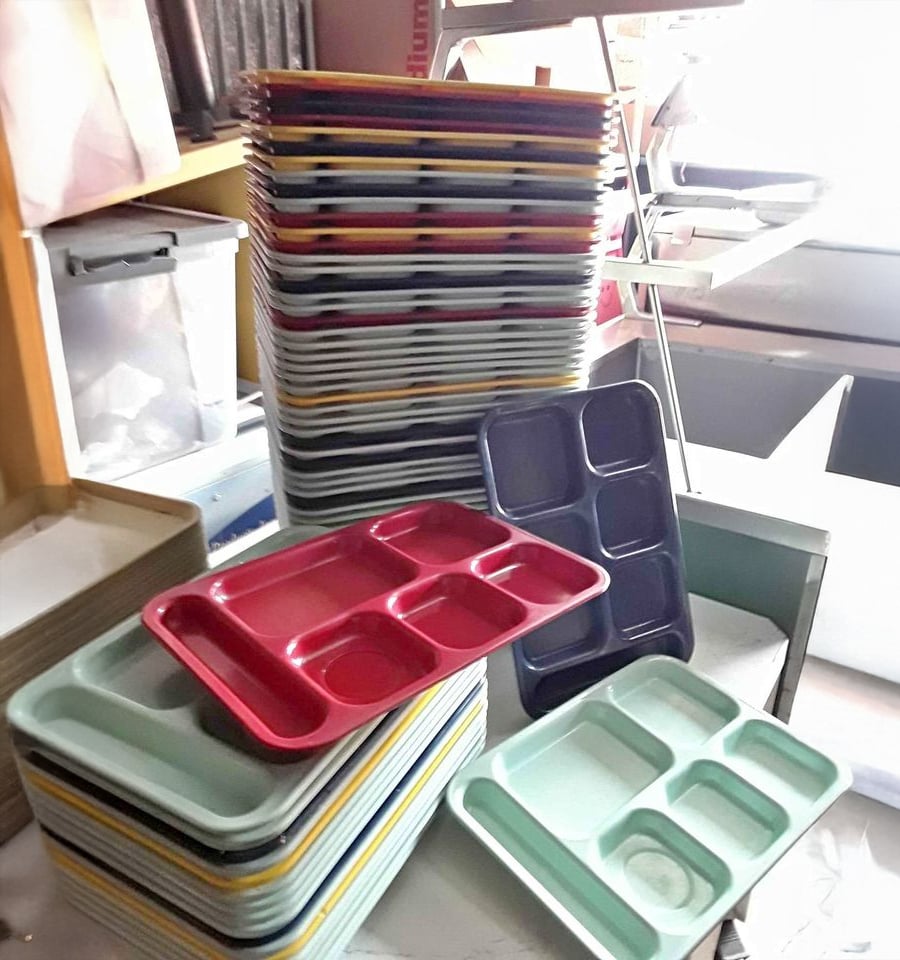 Stacks of colorful cafeteria trays, mostly rectangular with divided sections, are piled on a table and leaning against a shelf, with various trays in shades of red, green, yellow, black, and blue.