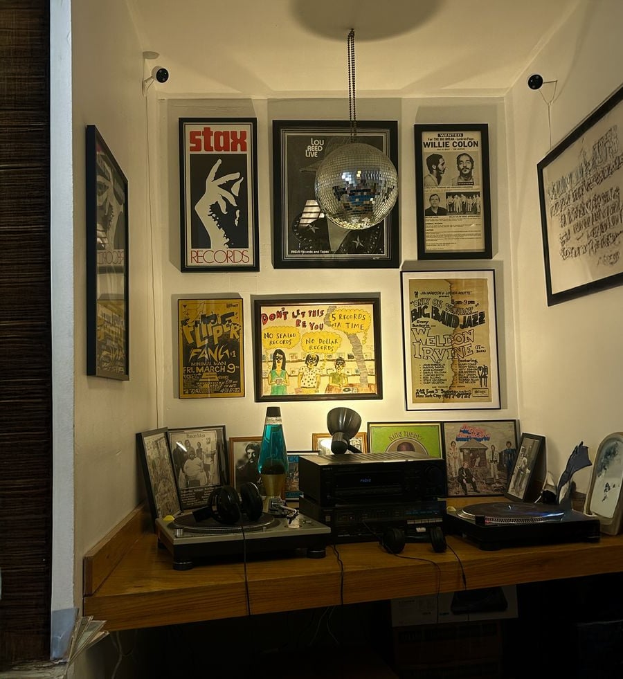A cozy corner with a wooden desk holding turntables, records, audio equipment, headphones, and a lava lamp. Framed vintage music posters decorate the walls, and a small disco ball hangs from the ceiling.